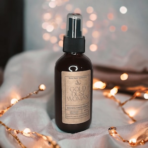 May include: A brown glass bottle with a black spray nozzle and a label that reads "Moon Water Apothecary Gold Dust Woman Room & Linen Spray 4 fl oz Infused with Moon Water Hand Poured in Cedar Rapids, IA"