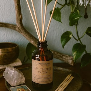 May include: A brown glass diffuser bottle with reed sticks, labeled "Patchouli Moon" by Moon Mamer. The bottle sits on a dark metal tray with a crystal and a tarot card. The background includes a plant and a decorative branch.