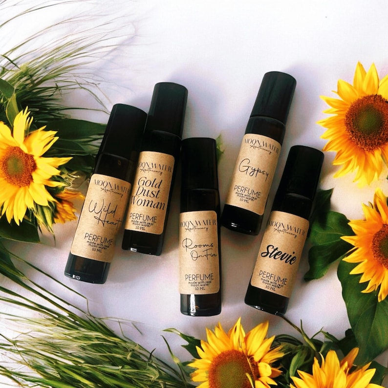 Pode incluir: Cinco pequenos frascos de vidro com r&oacute;tulos marrons e tampas pretas de rolagem. Os r&oacute;tulos t&ecirc;m o texto "Moonwater Apothecary" e os nomes de diferentes aromas: "Wild Heart", "Gold Dust Woman", "Rooms On Fire", "Gypsy" e "Stevie". Os frascos est&atilde;o dispostos sobre uma superf&iacute;cie branca com girass&oacute;is.