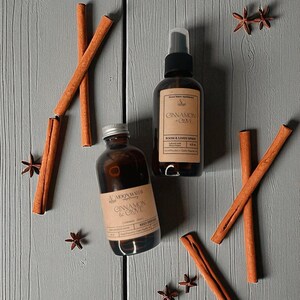 May include: Two brown glass bottles and cinnamon sticks are arranged on a gray wooden surface. One bottle is a room and linen spray, and the other is a reed diffuser. The labels read "Cinnamon & Clove". Star anise is scattered around the bottles.
