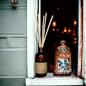 May include: A brown glass aromatherapy diffuser bottle with reed sticks and a decorative copper-toned jar. The diffuser bottle has a label that reads "GYPSY". The jars are placed on a white windowsill against a gray brick wall.