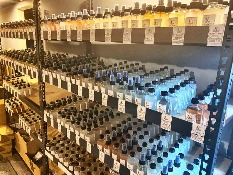 May include: A shelf unit filled with rows of small glass bottles with black lids and labels. The bottles are filled with a clear liquid and have labels with the text "Moon Water" and "Library".
