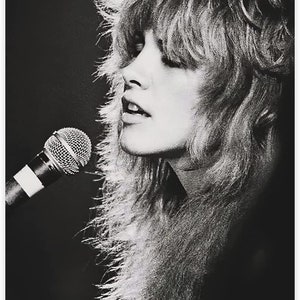 Pode incluir: Uma fotografia a preto e branco de uma mulher com cabelo comprido e em camadas a cantar num microfone. Ela est&aacute; a usar um colar de pele.