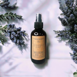 May include: A dark brown glass bottle of Lavender + Sage Room & Linen Spray. The label is a light brown with black text. The bottle is surrounded by sprigs of lavender and greenery. The spray bottle is 4 fl oz.