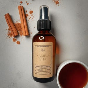May include: A brown glass bottle of Vanilla Chai room and linen spray with a black spray nozzle. The label reads "Vanilla Chai Room & Linen Spray". Cinnamon sticks and a cup of tea are also in the image.