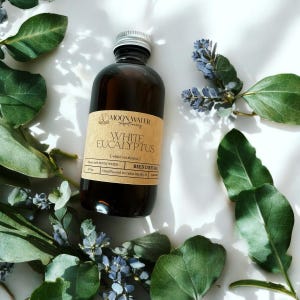 May include: A brown glass bottle with a white label that reads "MOON WATER Apothecary WHITE EUCALYPTUS [white eucalyptus] REED DIFFUSER Hand Poured in Cedar Rapids, IA". The bottle is surrounded by green leaves and blue flowers.
