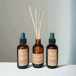 May include: Three amber glass bottles with black spray tops and brown paper labels. The bottles are labeled "Rosemary & Sage", "Pirake", and "Neroli" and are room and linen sprays. A brown glass bottle with a wooden diffuser stick set is also pictured. The bottles are on a white surface.