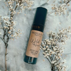Pode incluir: Um frasco de vidro marrom com um rolo e um r&oacute;tulo de papel marrom que diz "Wild Heart Perfume made with real moon water 10 ML". O frasco est&aacute; sobre um fundo de tecido azul claro e branco.