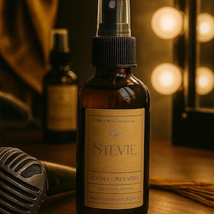 May include: A brown glass bottle of "Stevie" Boom & Linen Spray. The label reads "Moon Water Apothecary" and "Hand Poured in Cedar Rapids 34." The bottle has a black spray nozzle. The background includes a microphone and a blurred second bottle.
