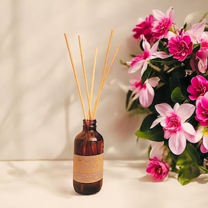 May include: A brown glass bottle with a brown label that reads "Moon Valley Plumeria Reed Diffuser". The bottle is filled with a clear liquid and has wooden sticks sticking out of it. The bottle is sitting on a white surface in front of a bouquet of pink flowers.