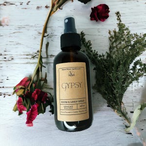 May include: A dark amber glass bottle of "Gypsy" room and linen spray. The label reads "Moon Water Apothecary" and "4 fl oz". The bottle is surrounded by dried flowers and greenery, creating a natural aesthetic.