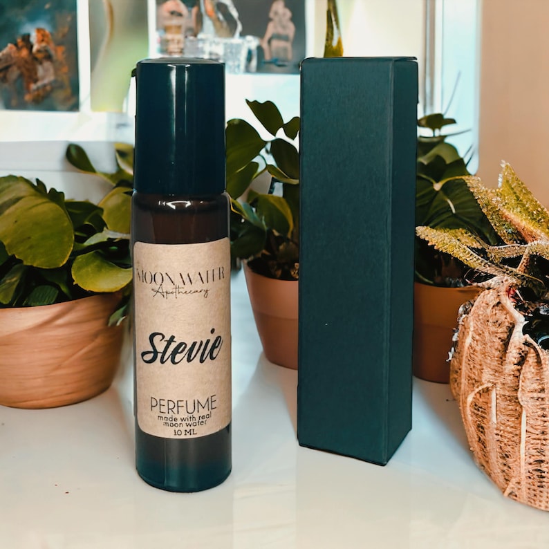 Pode incluir: Um frasco de vidro marrom com tampa preta e um r&oacute;tulo de papel marrom. O r&oacute;tulo diz "Moonwater Apothecary" e "Stevie Perfume" com o texto "made with real moon water 10 ML".
