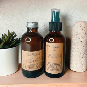 May include: Two amber glass bottles with brown labels. The bottle on the left is a 4 fl oz reed diffuser with the text "MOONWATER Apothecary GREEN TEA & LEMONGRASS | Fresh lemongrass, tea tree & bergamot | Infused with MOON WATER | Hand Poured in Cedar Rapids, IA". The bottle on the right is a 4 fl oz room and linen spray with the text "Moon Water Apothecary GREEN TEA & LEMONGRASS | ROOM & LINEN SPRAY | Infused with MOON WATER | Hand Poured in Cedar Rapids, IA".