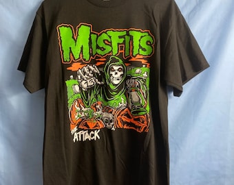May include: Black t-shirt featuring the word "MISFITS" in green and orange, with a graphic of a grim reaper holding a lantern and pumpkins. The text "THE ATTACK" is below the graphic.
