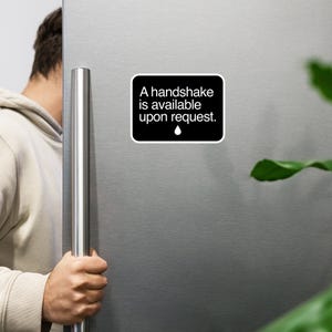 May include: A black and white sign on a stainless steel refrigerator that reads "A handshake is available upon request." A small water drop icon is below the text.