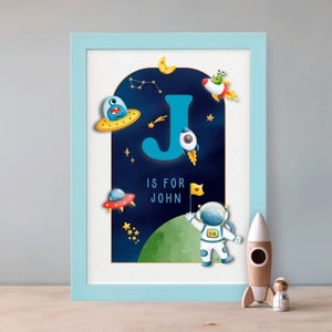 Puede incluir: Una impresión enmarcada azul y blanca con una letra J azul en una escena espacial con estrellas, un cohete, una nave espacial y un astronauta. El texto "J is for John" está debajo de la letra.