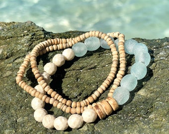Può includere: Una collana a strati con perline blu chiaro e beige. Le perline sono fatte di legno e vetro. La collana è su una superficie rocciosa.