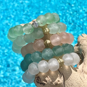 May include: A stack of bracelets made of light green, pink, and white glass beads. The beads are round and have a slightly irregular shape. The bracelets are stacked on top of each other, with the green beads at the bottom, the pink beads in the middle, and the white beads at the top.