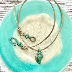 May include: A turquoise and gold seashell pendant necklace with a brown leather cord. The necklace is displayed on a turquoise and white ceramic plate.