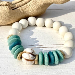May include: A white and teal beaded bracelet with a white and brown patterned bead in the center. The bracelet is made with round wooden beads and flat, teal and white beads.