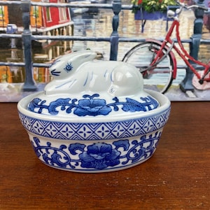 May include: A white and blue ceramic trinket box with a rabbit lid. The box features blue floral and geometric patterns. The lid is shaped like a rabbit. The box is on a wooden surface.