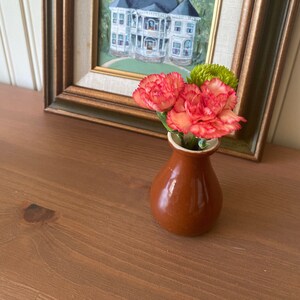 May include: A small brown ceramic vase with a white rim holds a bouquet of pink and green carnations. The vase is sitting on a wooden surface in front of a framed painting of a white house.