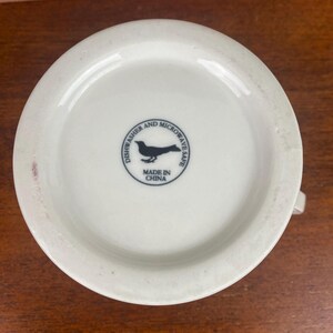 May include: A white ceramic mug base with a handle. The base has a black bird silhouette inside a circle with the words "Dishwasher and Microwave Safe" and "Made in China".