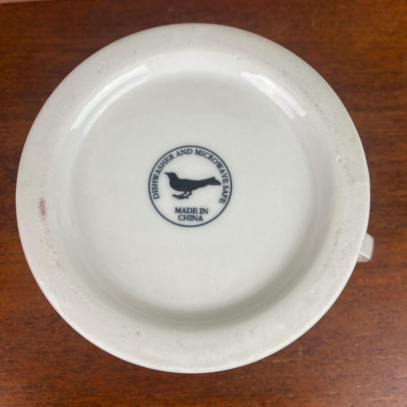May include: A white ceramic mug base with a handle. The base has a black bird silhouette inside a circle with the words "Dishwasher and Microwave Safe" and "Made in China".