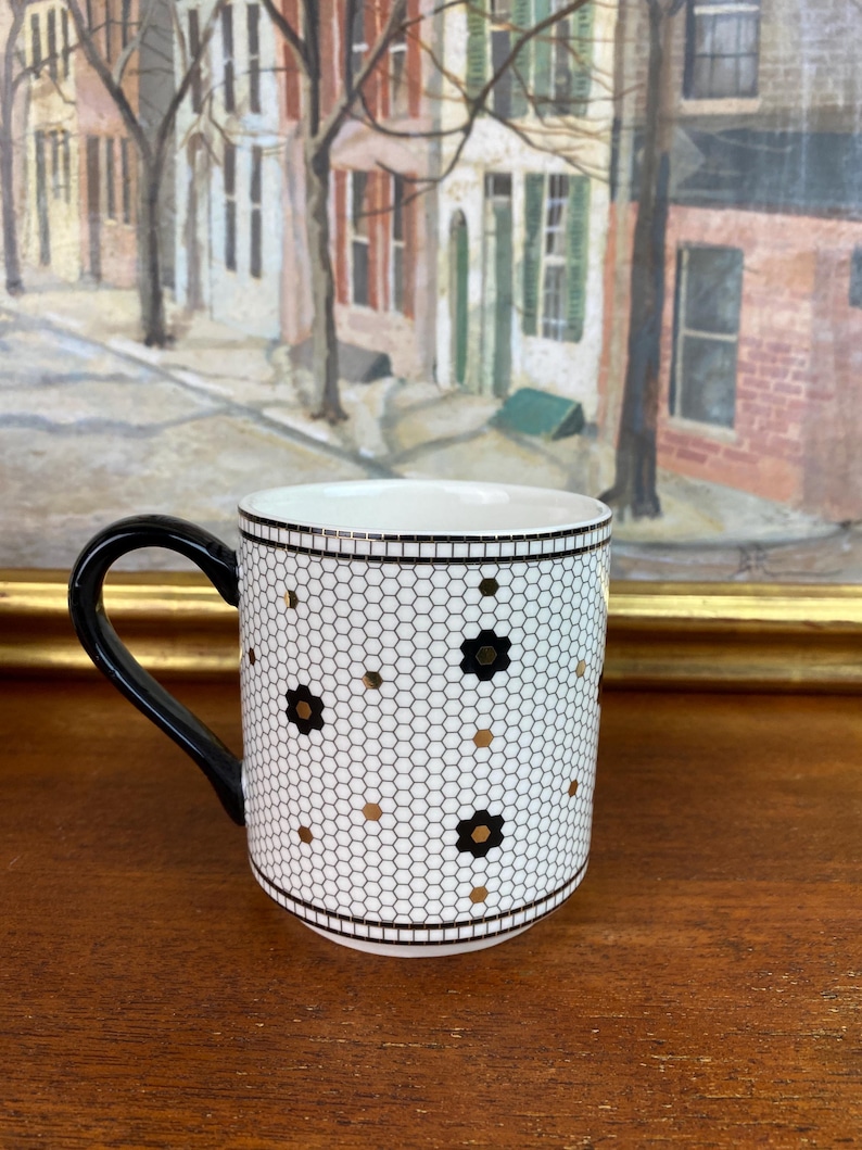 May include: White ceramic mug with a black handle. The mug has a black and gold geometric pattern that resembles a honeycomb.