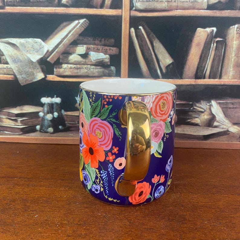 May include: A ceramic mug with a navy blue background, adorned with a vibrant floral pattern in shades of pink, orange, and yellow. The mug features a gold-colored handle and rim, adding a touch of elegance to the design.