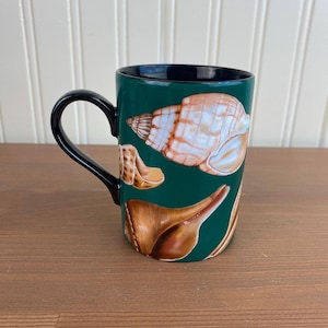May include: A ceramic mug with a dark green exterior and a black handle and interior. The mug features illustrations of various seashells in shades of brown, tan, and white. The mug is sitting on a wooden surface.