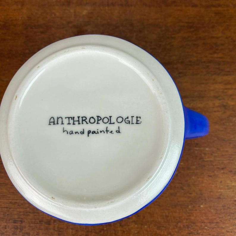 May include: A white ceramic mug with a blue handle and rim. The base of the mug features the text "ANTHROPOLOGIE hand painted" in black lettering. The mug is set against a wooden surface.