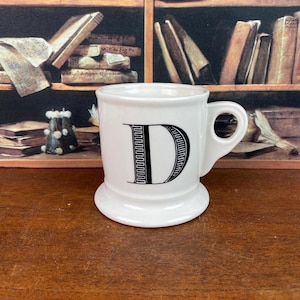 May include: White ceramic mug with a large black letter "D" on the front. The mug has a small handle and a wide base. The background features a bookshelf with books.