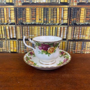 May include: A white teacup and saucer set with a floral design featuring roses in shades of red, pink, and yellow. The cup has a gold handle and trim. The saucer also has a gold trim. The set is on a wooden surface.