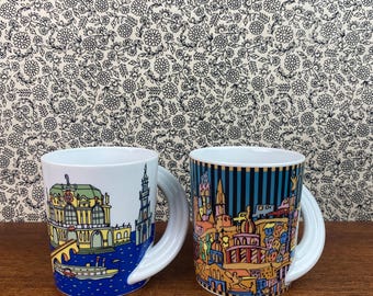 May include: Two white ceramic mugs with unique handles. The mugs feature colourful illustrations of cityscapes. One mug depicts a cityscape with a river and boats, while the other mug shows a cityscape with buildings and a car.