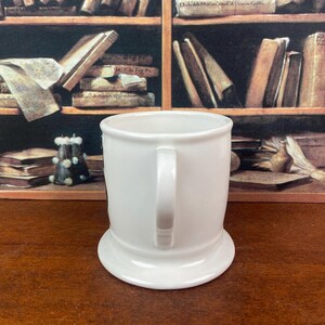 May include: A white ceramic mug with a handle and a wide, stable base. The mug has a simple, classic design, perfect for coffee or tea. The background features a shelf with books.