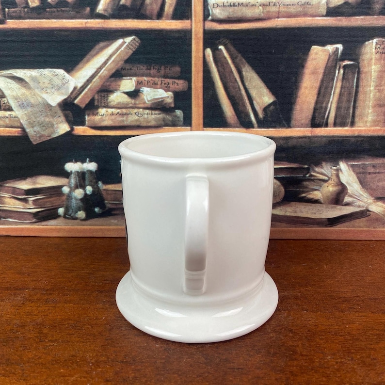 May include: A white ceramic mug with a handle and a wide, stable base. The mug has a simple, classic design, perfect for coffee or tea. The background features a shelf with books.