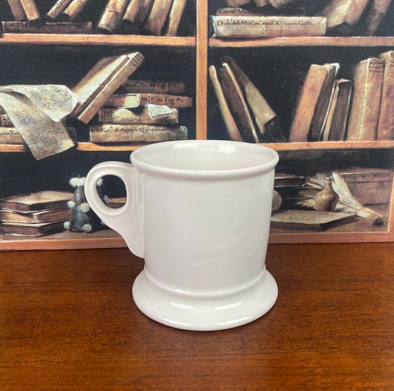 May include: A white ceramic shaving mug with a handle and a wide base. The mug has a classic design. It is set against a backdrop of a bookshelf with old books.
