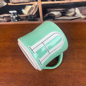May include: A mint green ceramic mug with a white letter "P" design. The mug has a curved handle and a white interior. The mug is on a wooden surface.