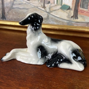 以下が含まれることがあります： 横たわった姿勢のボルゾイ犬の光沢のある陶器製フィギュア。この彫刻は、白いベースコートに、頭部、耳、背中、後肢を覆う特徴的な黒い斑点模様が施されており、滑らかで反射性のある釉薬仕上げが特徴です。
