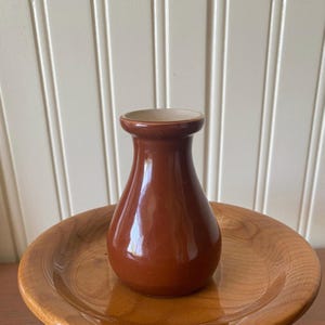 May include: A small, glossy, dark brown ceramic vase with a rounded body and a flared rim, centered on a smooth, light brown wooden plate. The vase has a smooth, reflective surface and a simple, functional shape with a narrow neck.