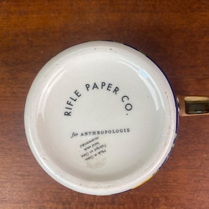 May include: White ceramic mug with a colorful floral design and a gold handle. The bottom of the mug features the text "RIFLE PAPER CO." and "for ANTHROPOLOGIE". Hand wash recommended.