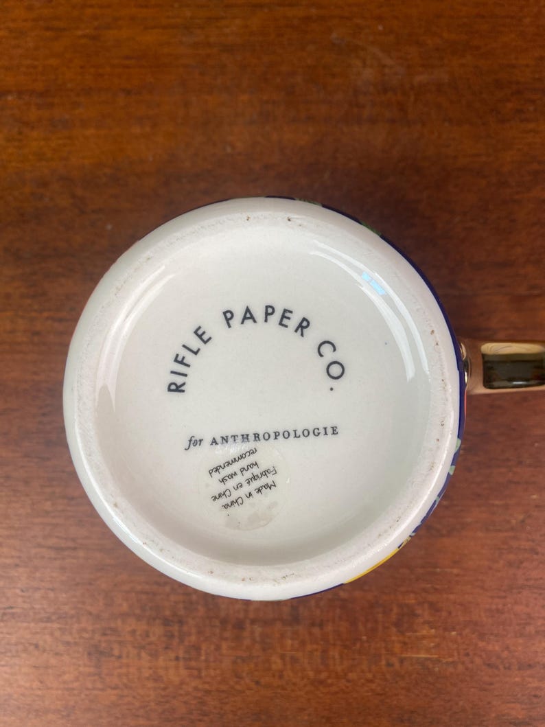 May include: White ceramic mug with a colorful floral design and a gold handle. The bottom of the mug features the text "RIFLE PAPER CO." and "for ANTHROPOLOGIE". Hand wash recommended.