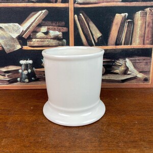 May include: A white ceramic tumbler with a rounded top and a flared base. The tumbler is cylindrical and has a smooth, glossy finish. The base is approximately 7.6 cm in diameter.