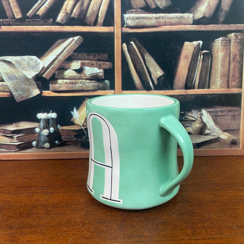 May include: A mint green ceramic mug with a white interior and a large white letter "A" on the front. The mug has a curved handle and sits on a wooden surface. The background features a painting of a bookshelf.