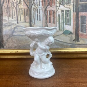 Könnte beinhalten: Weiße Keramikfigur eines Cherubins, der eine Schale hält. Der Cherub steht auf einem Sockel mit floralem Design. Die Schale ist leer.