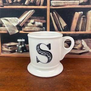 May include: White ceramic mug with a large, ornate black letter "S" on the front. The mug has a rounded base and a handle. The background features a bookshelf with books.