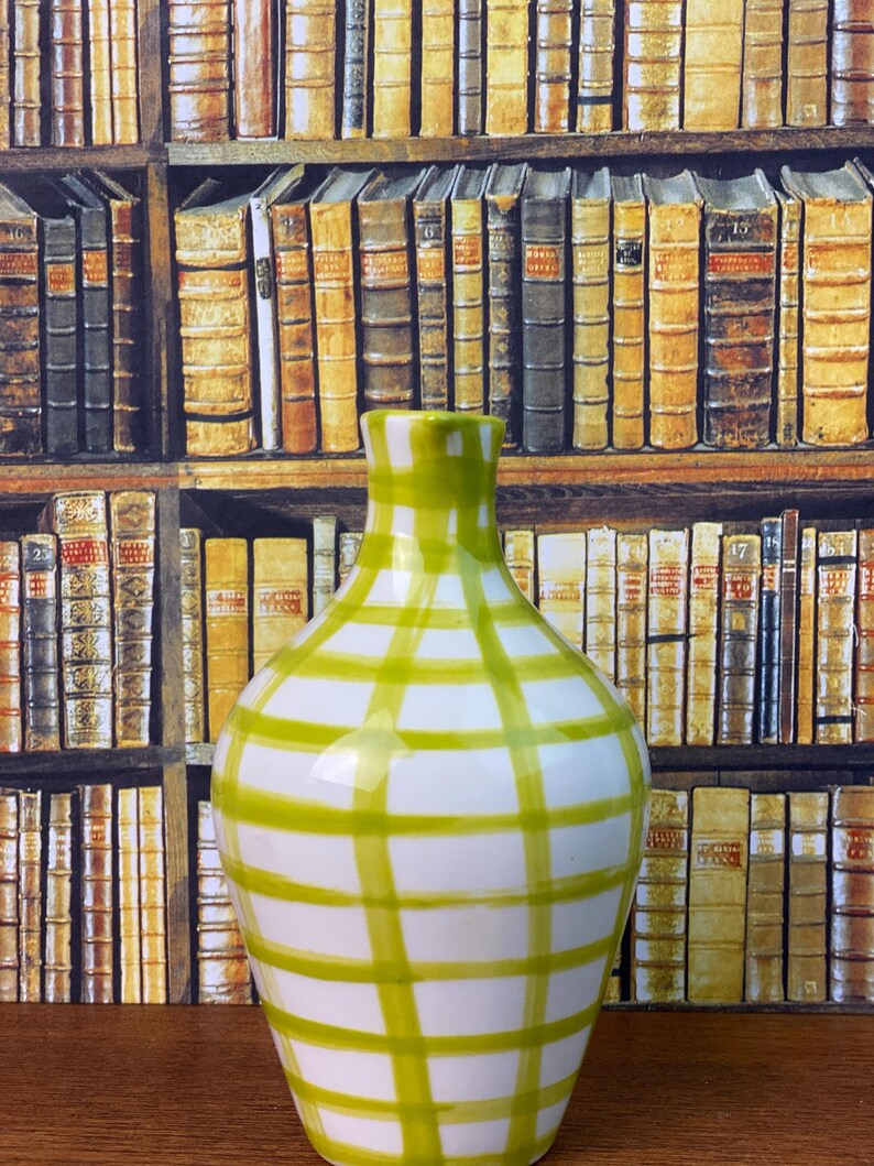 May include: A white ceramic vase with a green grid pattern. The vase has a rounded body and a narrow neck. The background shows a wallpaper design of bookshelves filled with books.