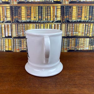 May include: A white ceramic mug with a curved handle. The mug has a wide base and a slightly flared rim. A black graphic is visible on the side of the mug. The background features a wall of books.