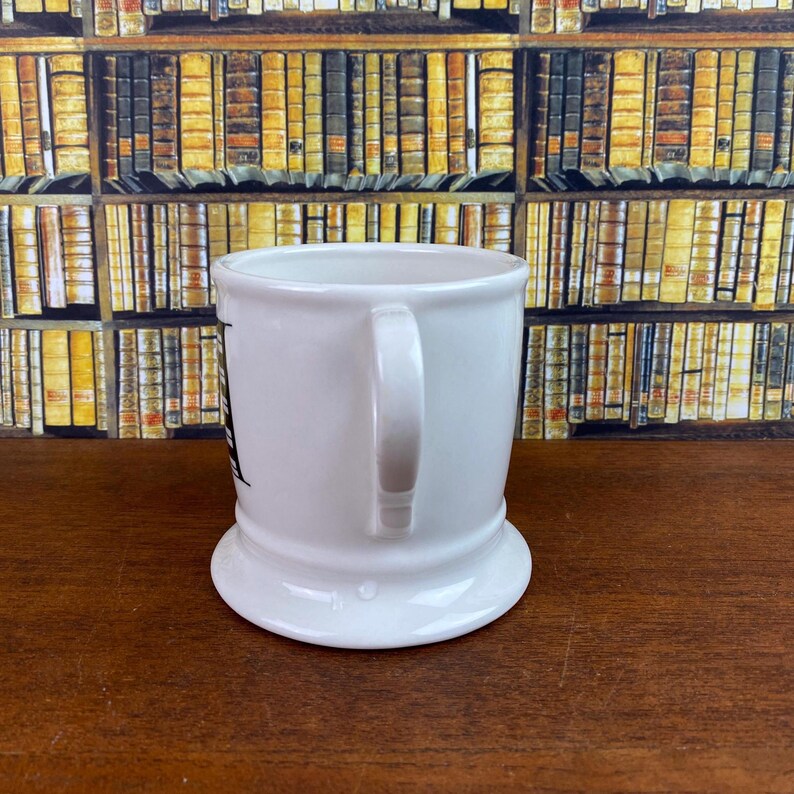 May include: A white ceramic mug with a curved handle. The mug has a wide base and a slightly flared rim. A black graphic is visible on the side of the mug. The background features a wall of books.